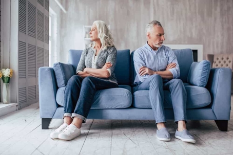 A seniors couple sitting on opposite ends of a couch with their backs to each other.